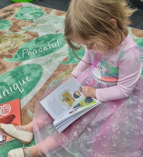 A little girl reading a book. She's wearing a pink fairy outfit.