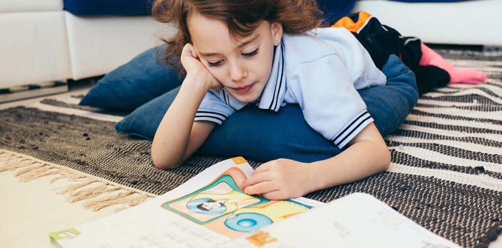 A boy lounging on the floor, reading a picture book. 
